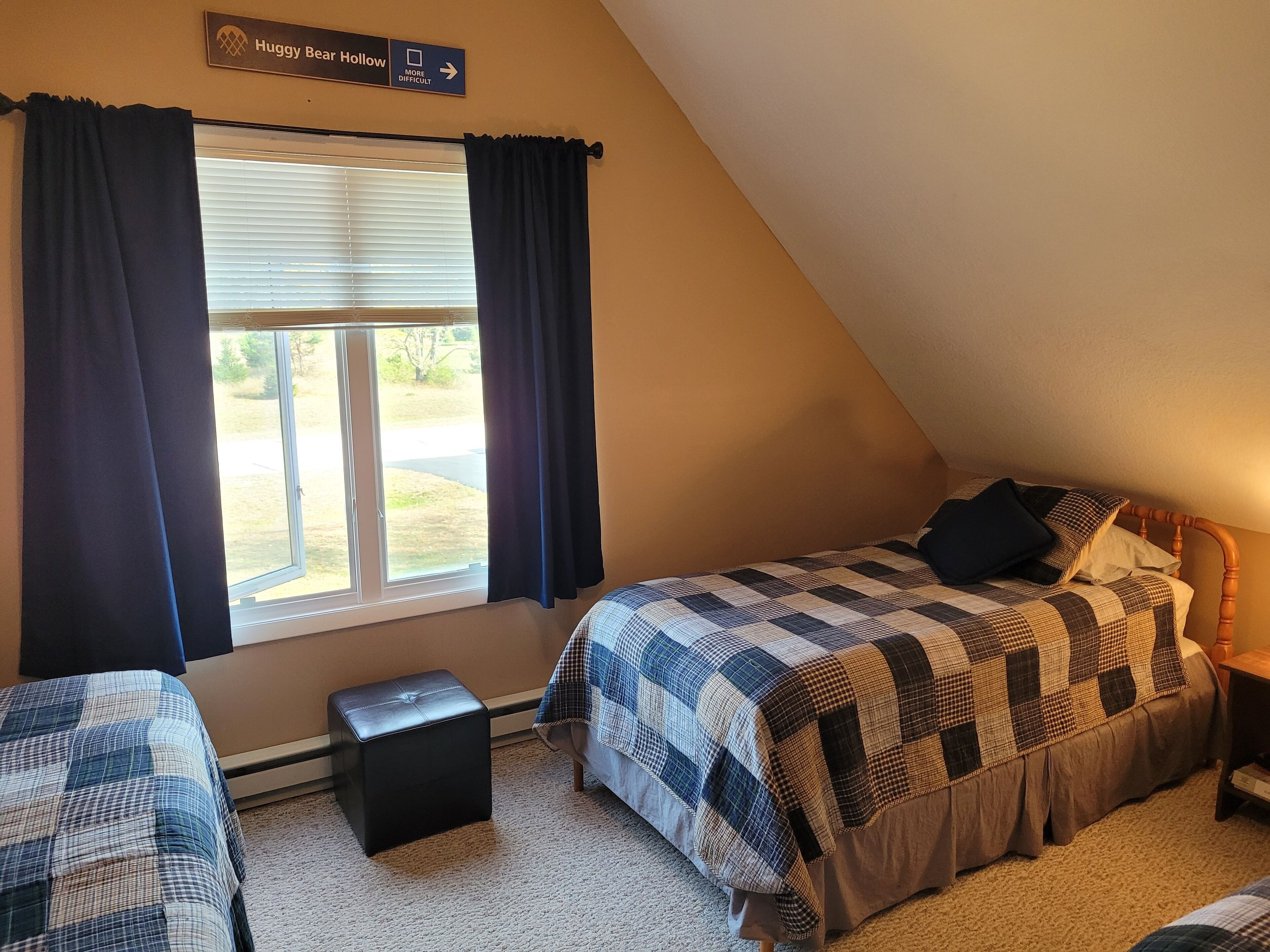 Un dormitorio con techo inclinado, una ventana con cortinas, una cama con colcha a cuadros, una mesita de noche y un taburete pequeño.