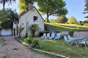 Une maison en pierre avec une échelle, un chemin de gravier et une zone herbeuse avec des chaises blanches et une table.