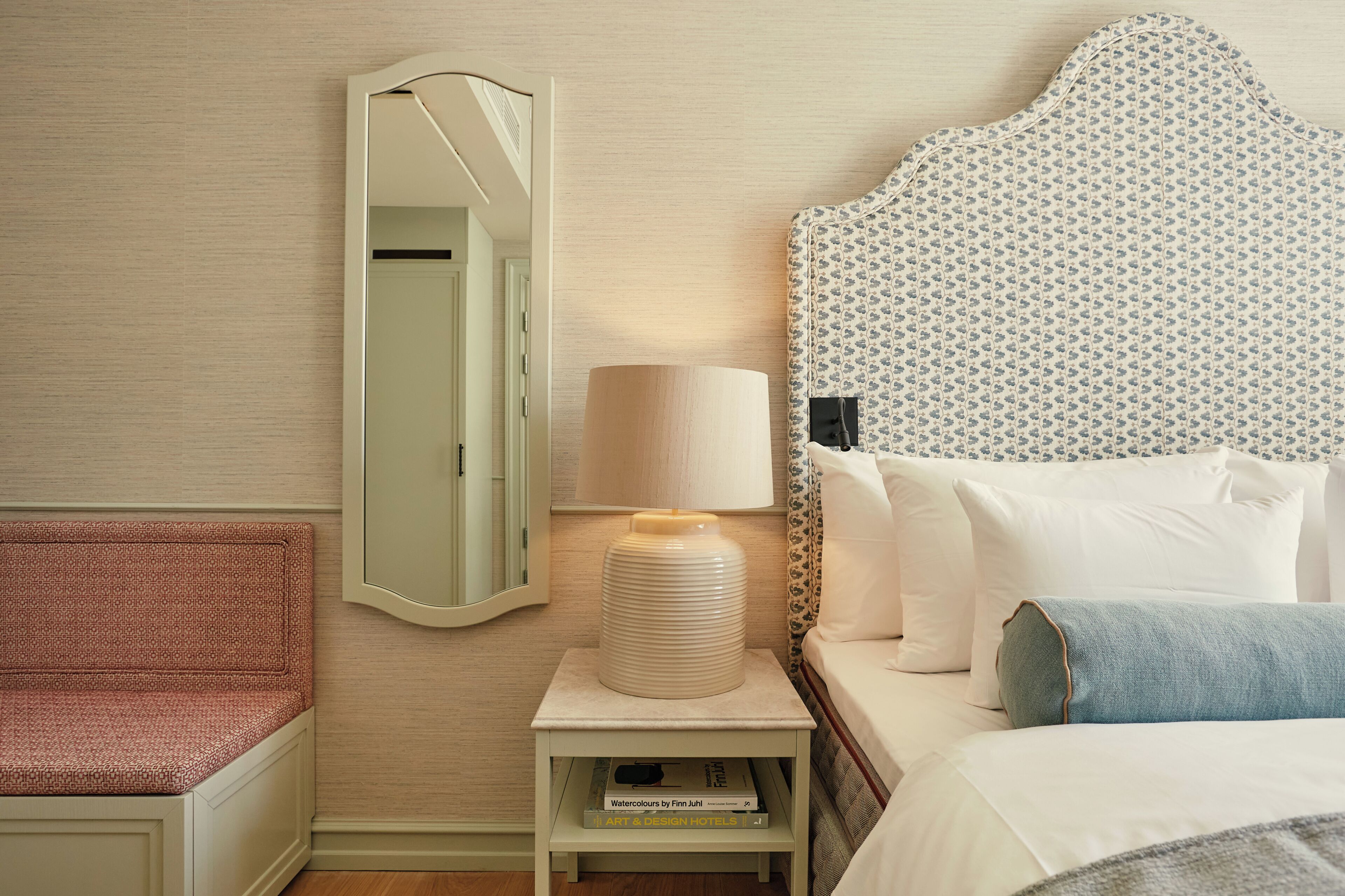 A hotel room with a bed, a bedside table with a lamp, a mirror, and a small shelf with books.