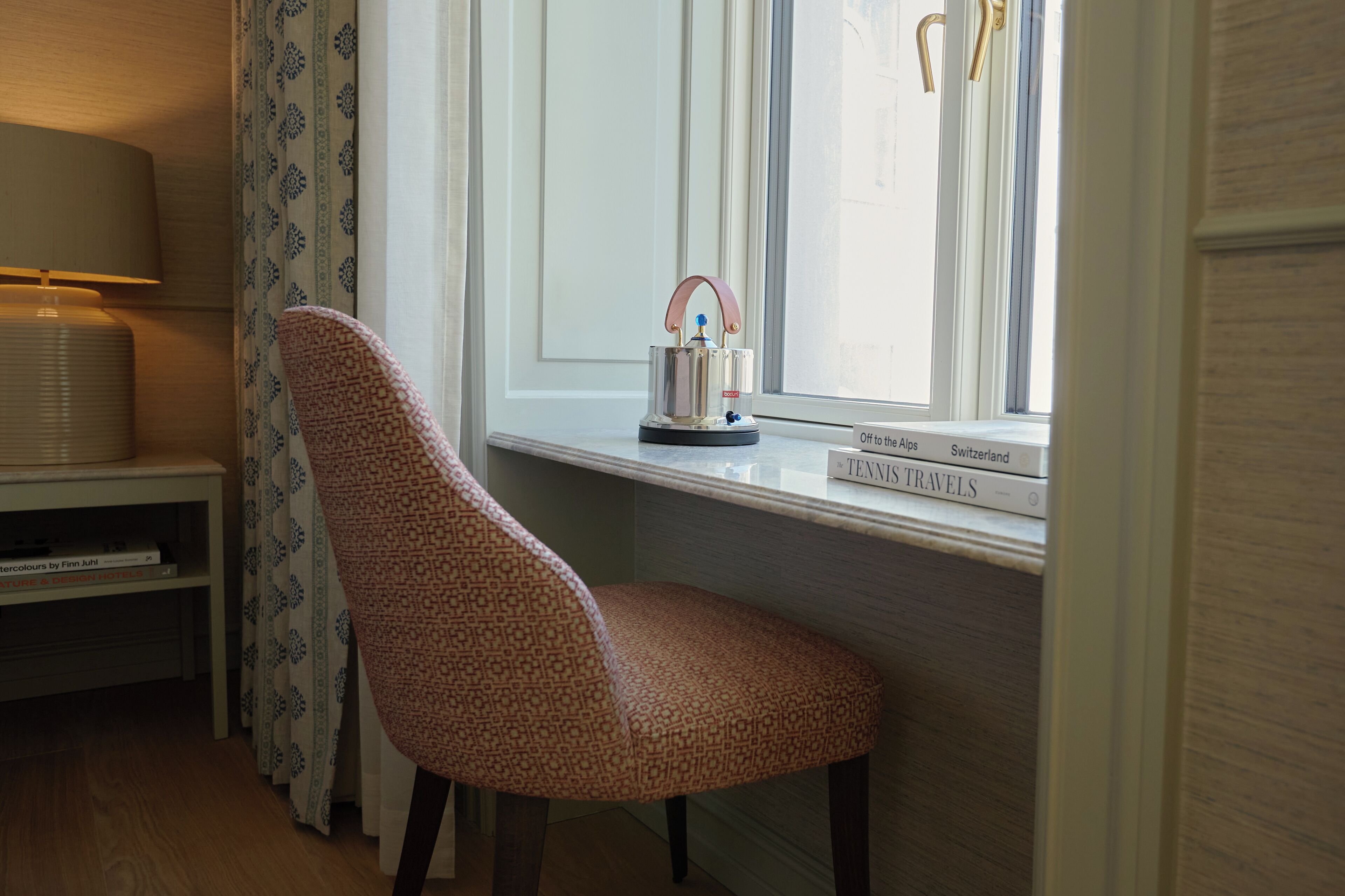 Una silla cerca de una ventana, con una lámpara sobre una mesita auxiliar, un libro titulado 'TENNIS TRAVELS' y una ventana que ofrece vistas al cielo.