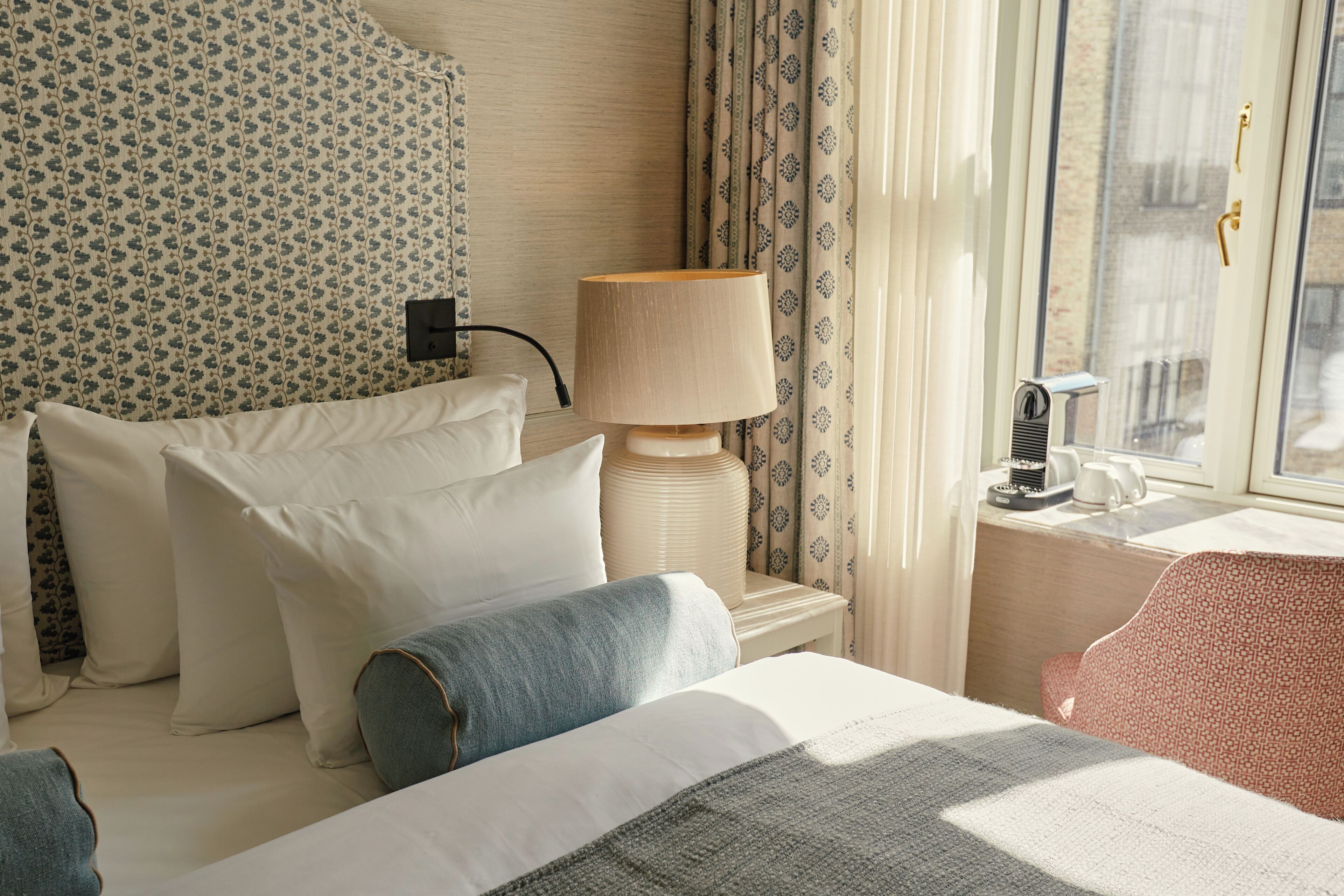 A hotel room with a bed, pillows, a lamp, a window with curtains, and a bedside table with a kettle.