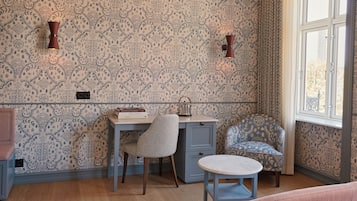 A room with patterned wallpaper, a desk with books, a chair, a small table, and a window with a view.