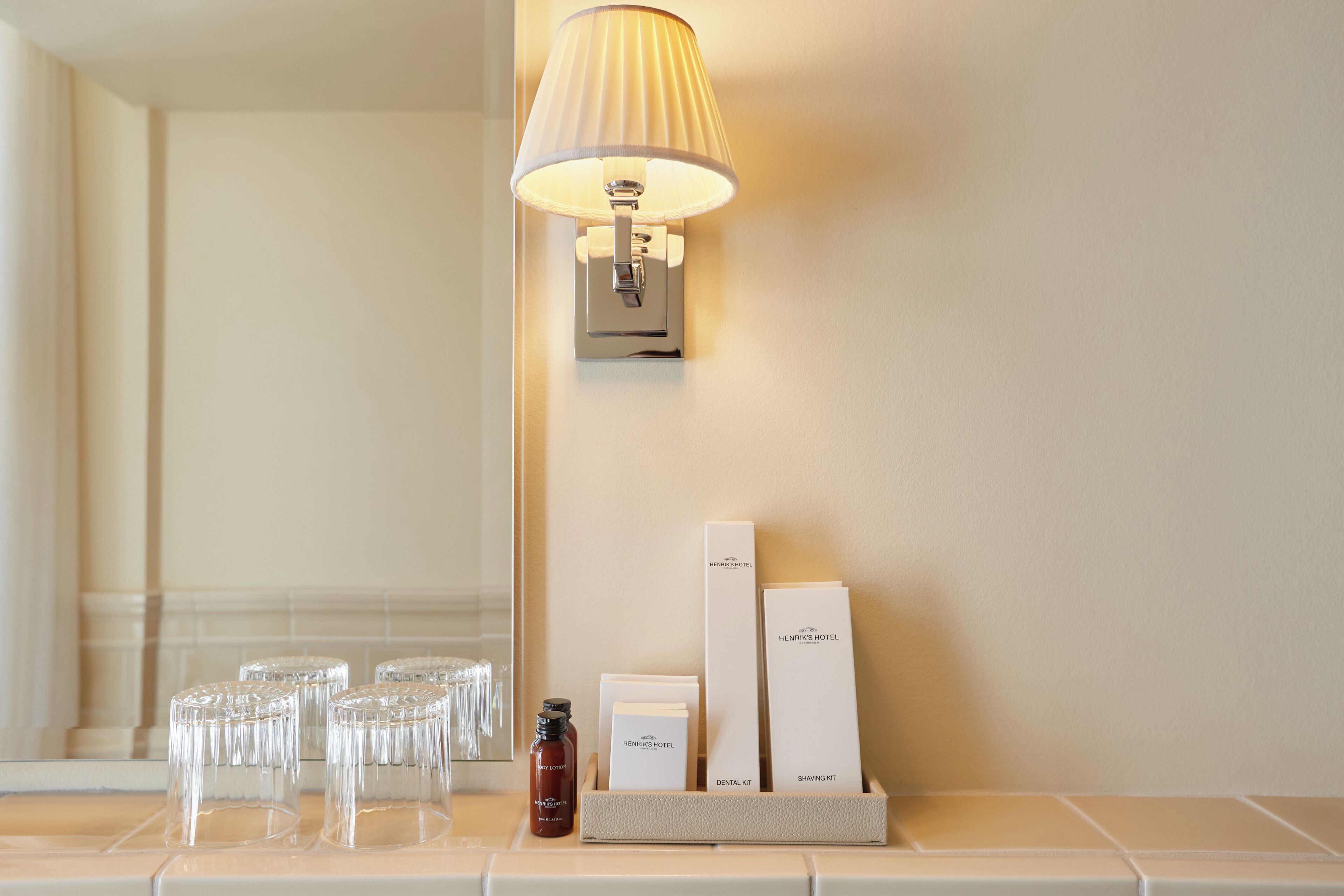 Un baño de hotel con espejo, una lámpara de pared y artículos de aseo que incluyen un cepillo de dientes, pasta dental y una maquinilla.