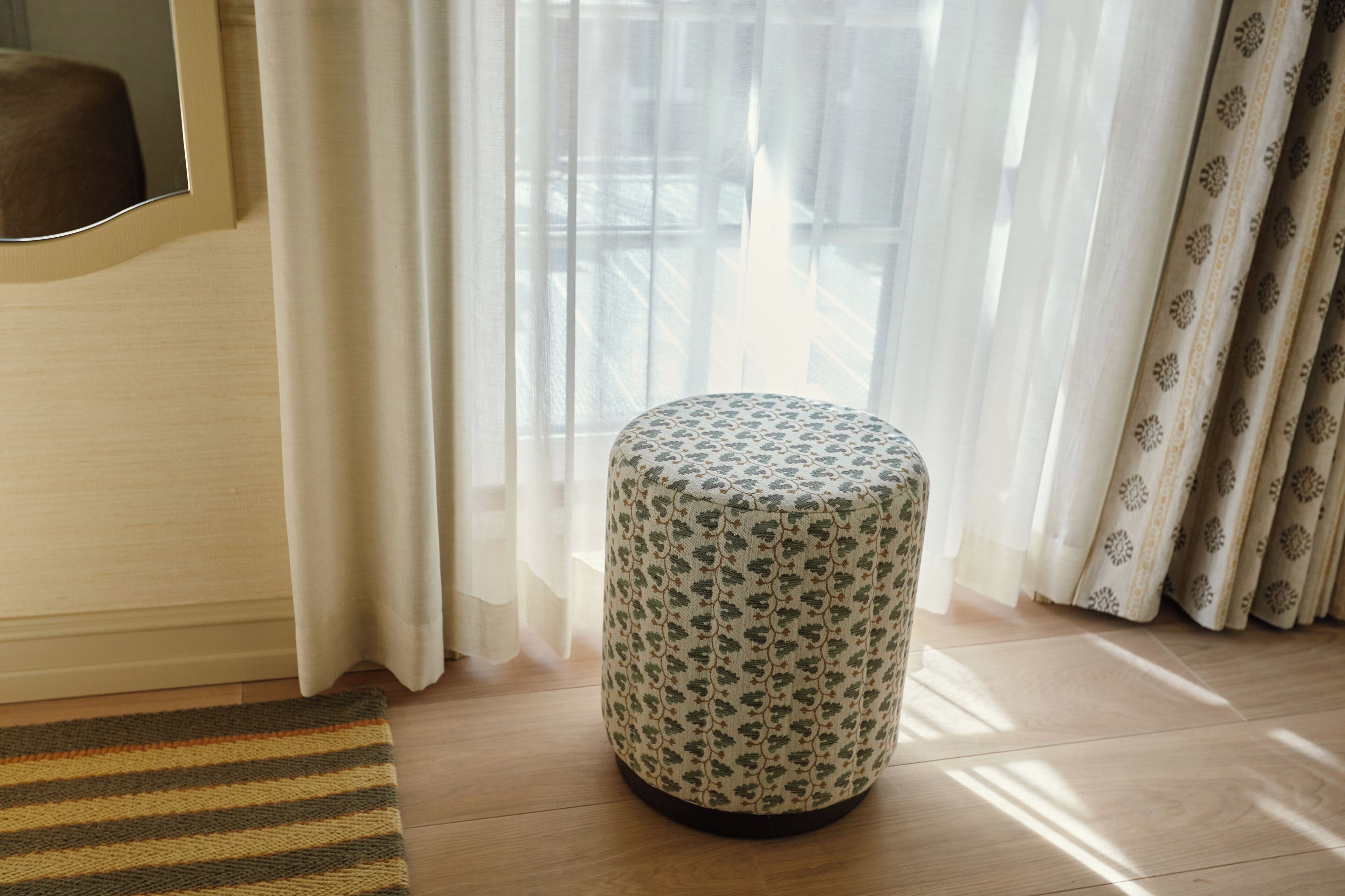 Una habitación con un otoman redondo, un tapete a rayas y cortinas traslúcidas.