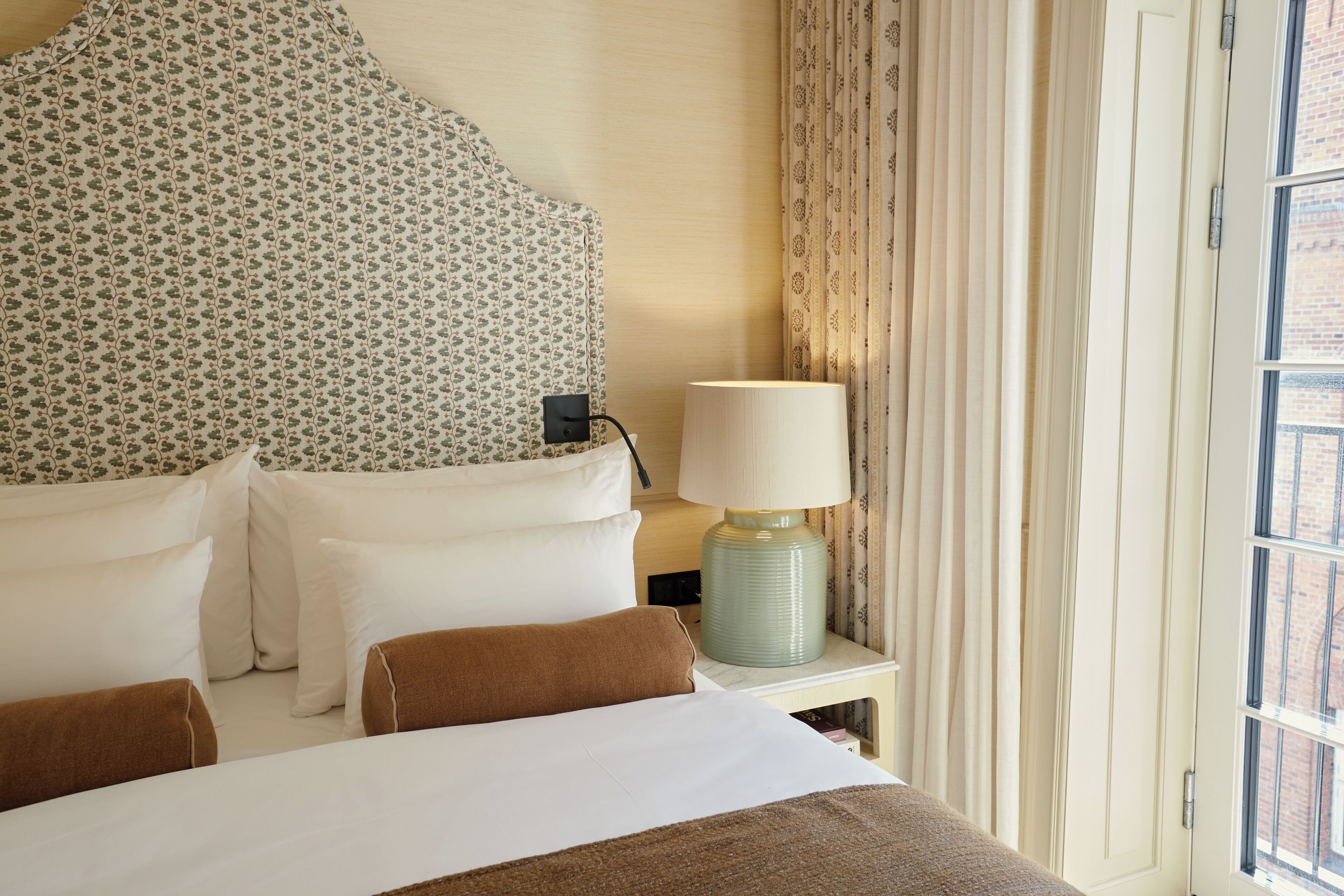 A hotel room with a bed, pillows, a lamp, and a window with curtains.