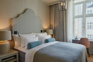 Habitación de hotel con una cama grande, cabecera con diseño estampado, dos lámparas de noche, una mesita con lámpara, una silla y una ventana con cortinas.