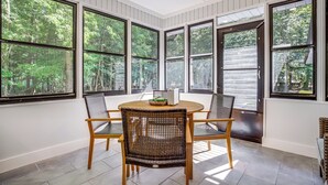 Un coin repas avec une table ronde et quatre chaises en osier, de grandes fenêtres donnant sur la verdure et une porte à cadre sombre.