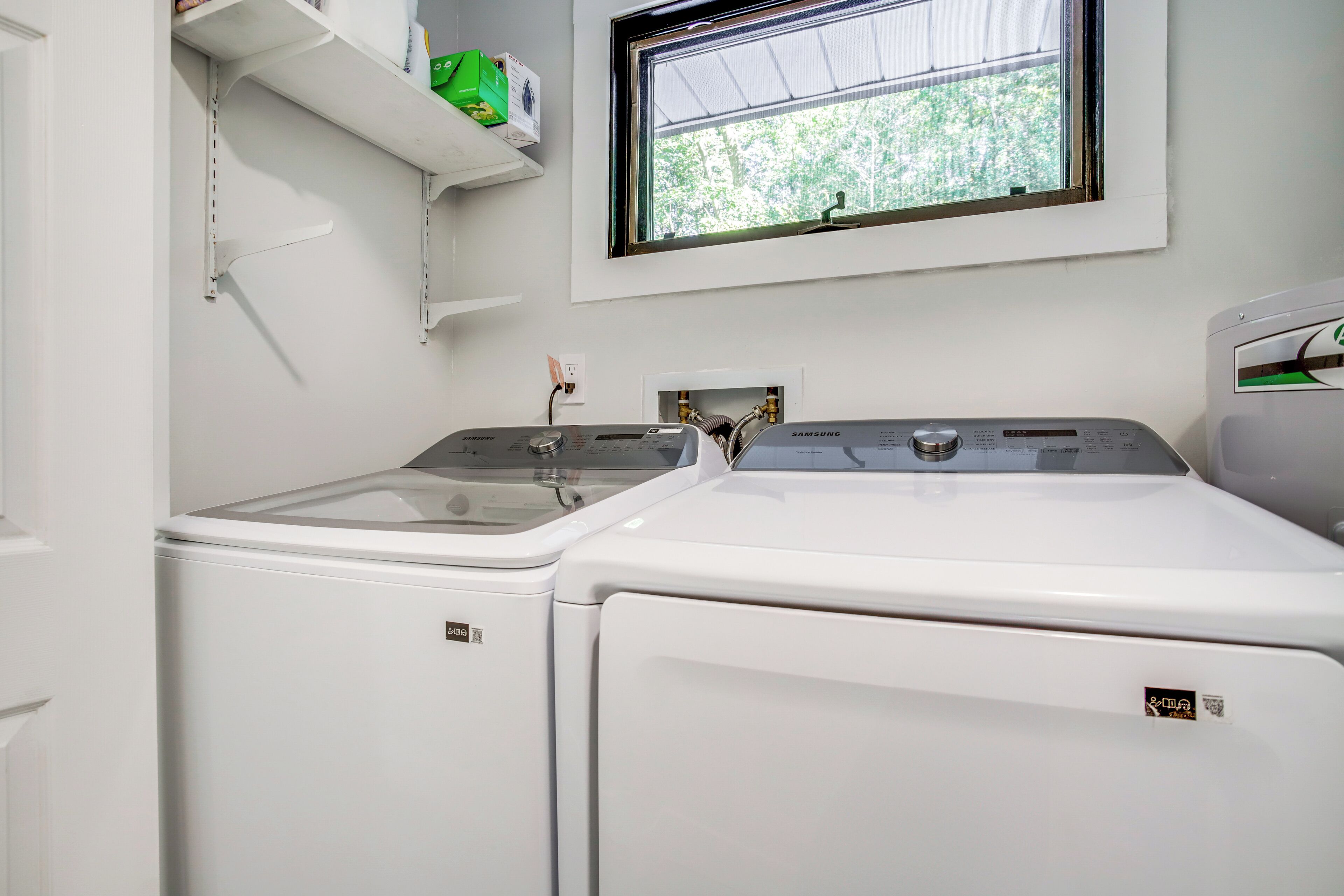 A laundry room with two white front-loading washing machines, a shelf with boxes, and a window with a view of trees.