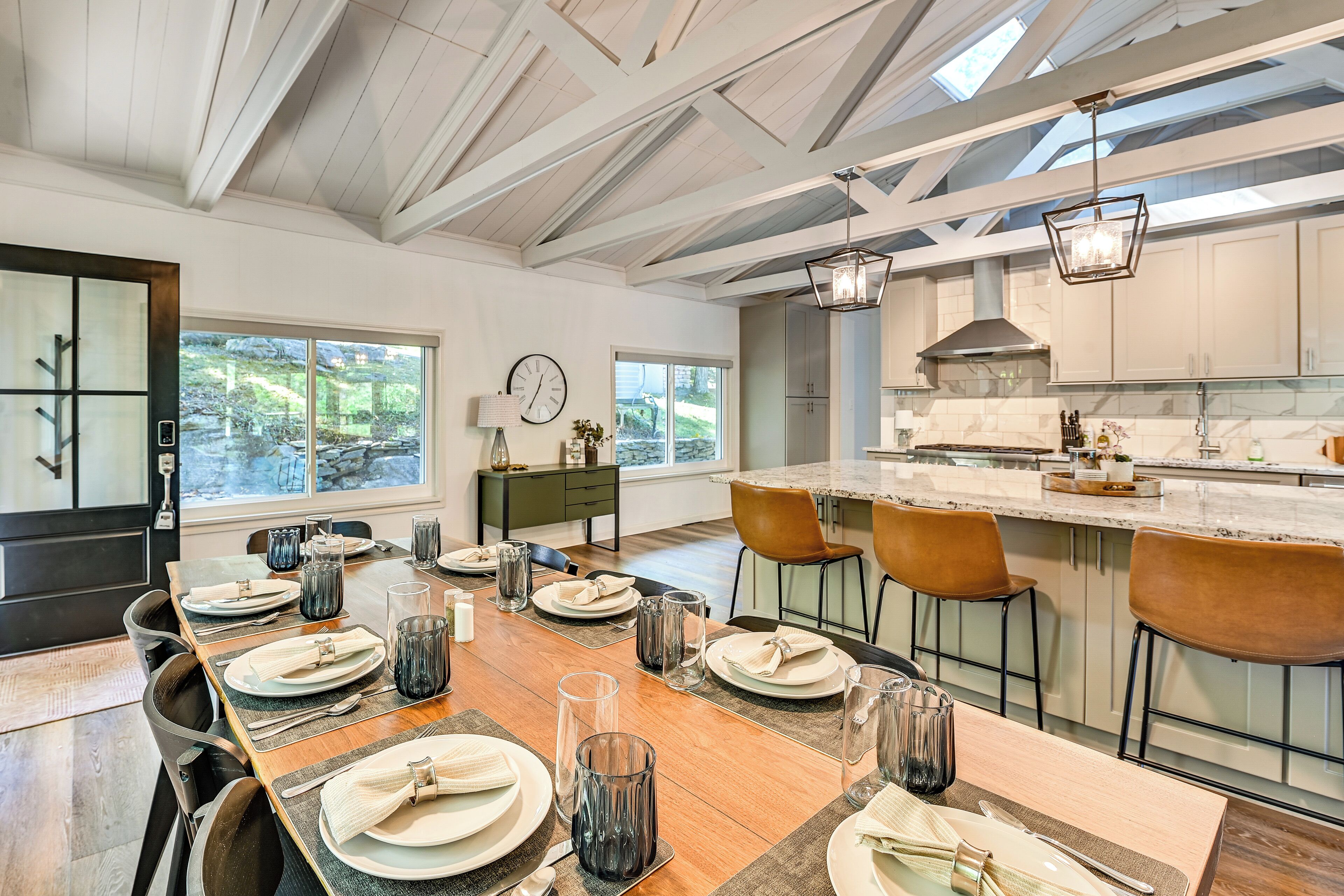 A modern kitchen with a large island, wooden dining table set for six, and a view of the outdoors through large windows.