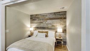 Une chambre à coucher avec une tête de lit en bois, un lit recouvert d’une literie blanche, deux tables de chevet avec des lampes et une petite table de nuit.