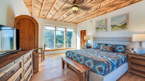 A bedroom with a wooden ceiling, large windows, a bed with patterned bedding, a TV, and a bench. - Panoramic Vistas: Glorieta Adobe Near Pecos River (Glorieta)