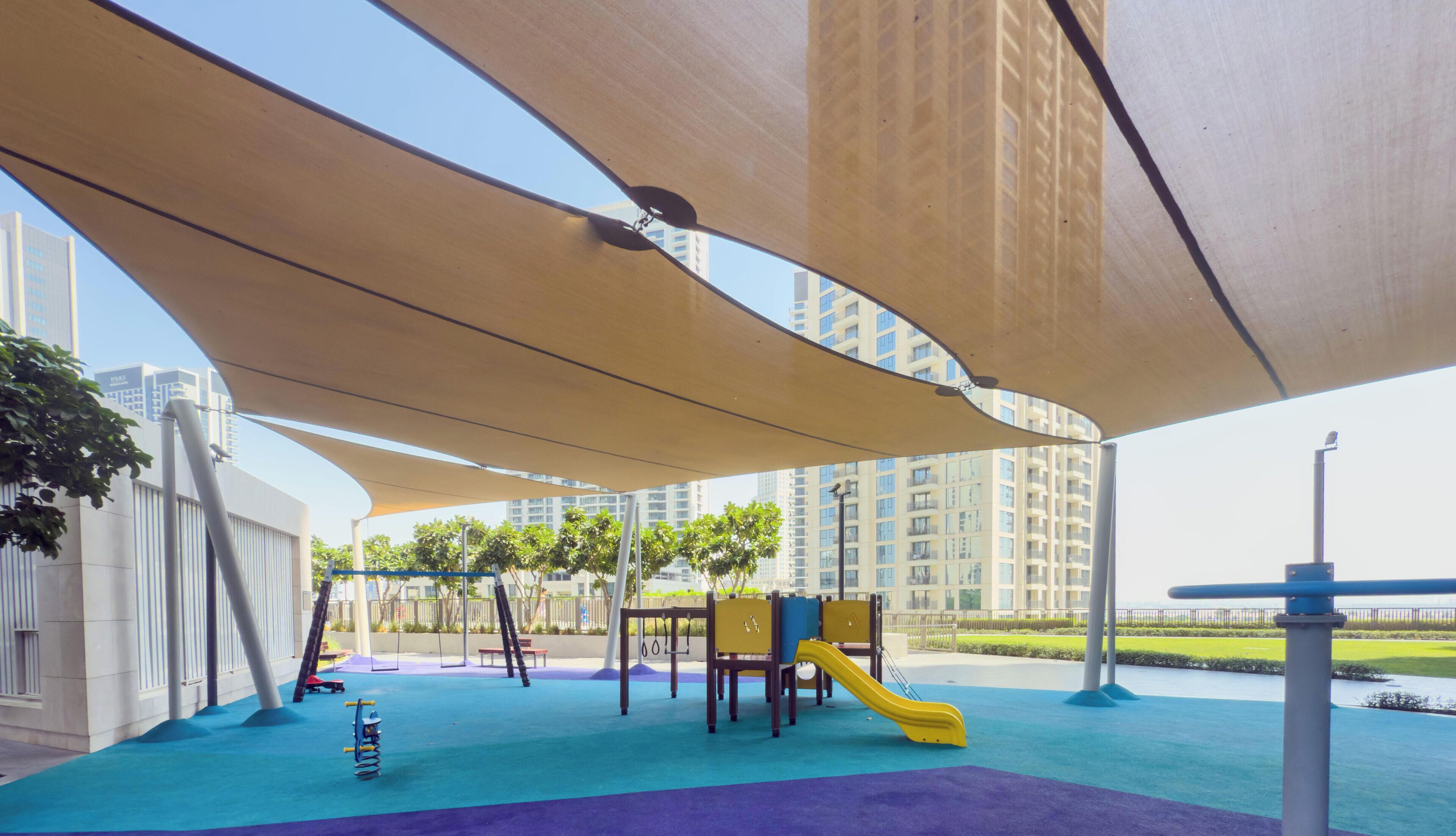 Un espace de jeux extérieur doté d’un toboggan et de structures d’escalade, ombragé par une grande marquise et entouré d’immeubles de grande hauteur.