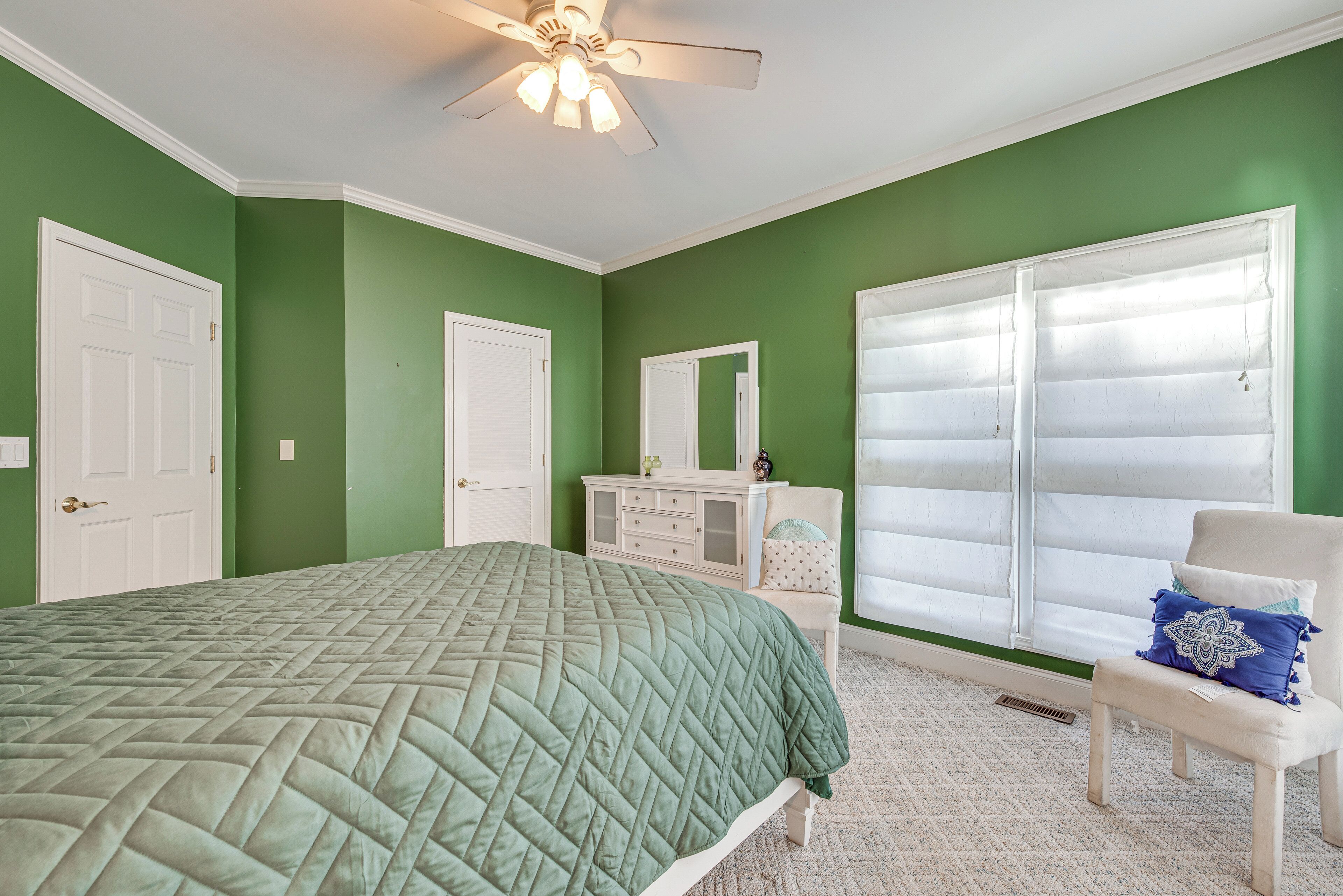Un dormitorio con pared verde, muebles blancos, una cama con colcha verde, un aparador blanco, una silla blanca y un ventanal con persianas.