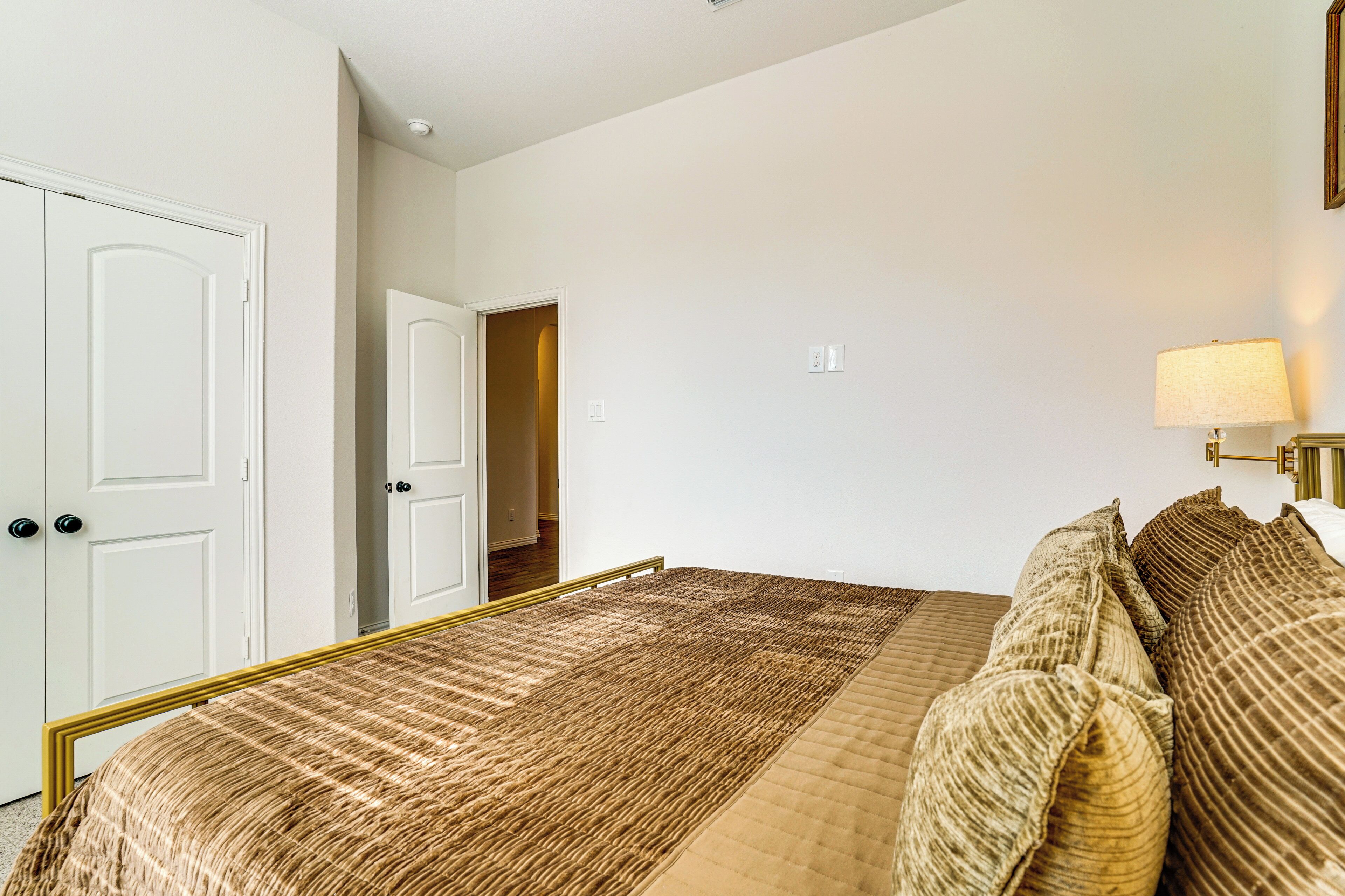 Um quarto com cama, mesa de cabeceira, abajur e porta para outro cômodo.