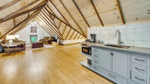 Un espacioso dormitorio en ático con techo inclinado, vigas de madera, una kitchenette integrada y una zona de estar acogedora con sofá y reposapiés.