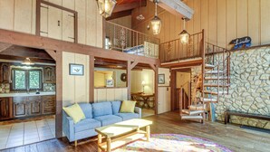 Ein großzügiger Wohnbereich mit einem blauen Sofa, einem Holztisch und einem farbenfrohen Teppich. Im Hintergrund sind eine Steinwand und eine Holztreppe zu sehen.