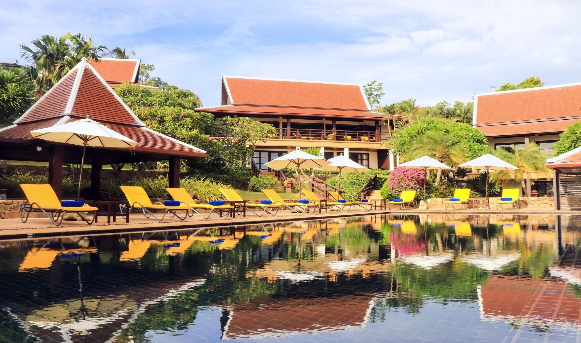 A poolside area with sun loungers and umbrellas, surrounded by buildings with red roofs.
