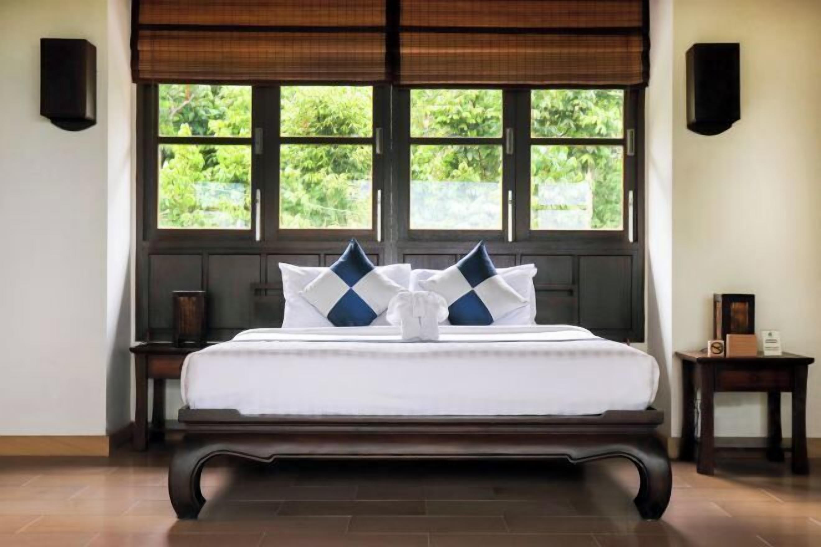 A neatly made bed with a white and blue checkered pillow arrangement, positioned in front of a window with a view of greenery.