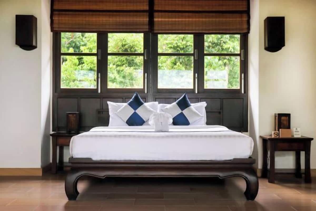 A neatly made bed with a white and blue checkered pillow arrangement, positioned in front of a window with a view of greenery.