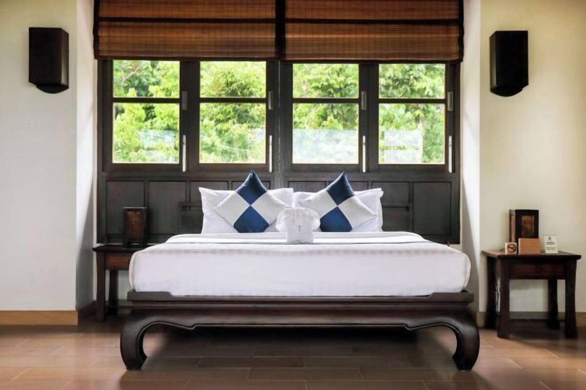 A neatly made bed with a white and blue checkered pillow arrangement, positioned in front of a window with a view of greenery.
