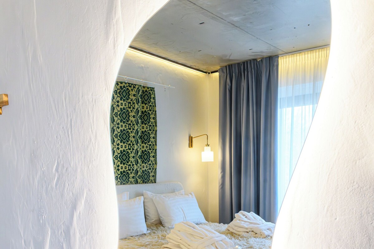 Una ventana circular que muestra un dormitorio con una cama, almohadas y un papel tapiz estampado.