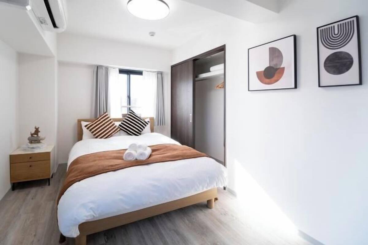 A modern hotel room with a bed, a wardrobe, a nightstand, and two framed artworks on the wall.