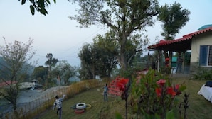 A grassy outdoor area with a fence, a tree, and a building with a red roof. - Dream Stay near Kainchi DHAM (Nainital)