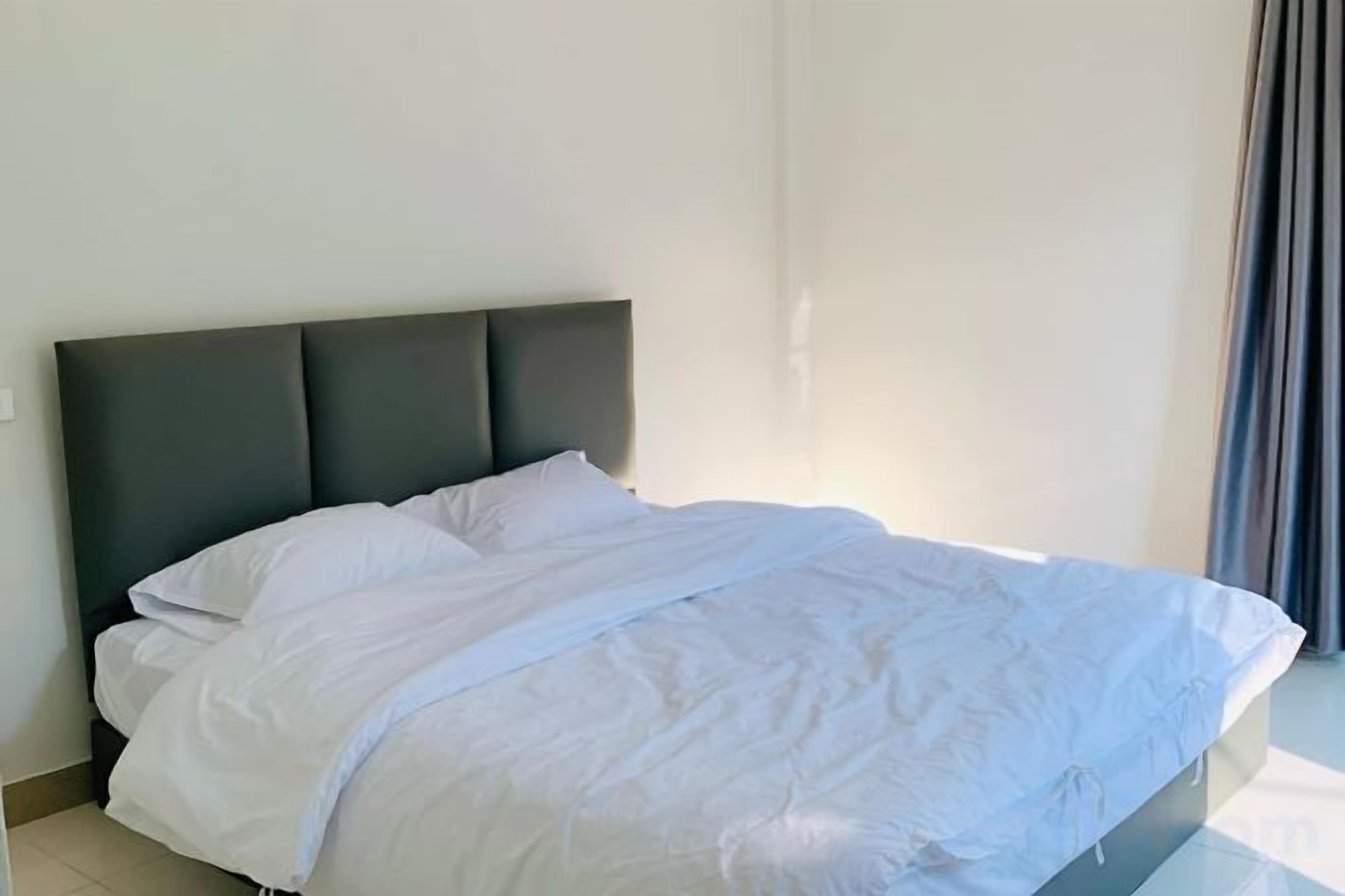 Un lit avec une tête de lit sombre, dans une pièce aux murs unis et dotée d’une fenêtre avec des rideaux.