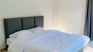 Un lit avec une tête de lit sombre, dans une pièce aux murs unis et dotée d’une fenêtre avec des rideaux.