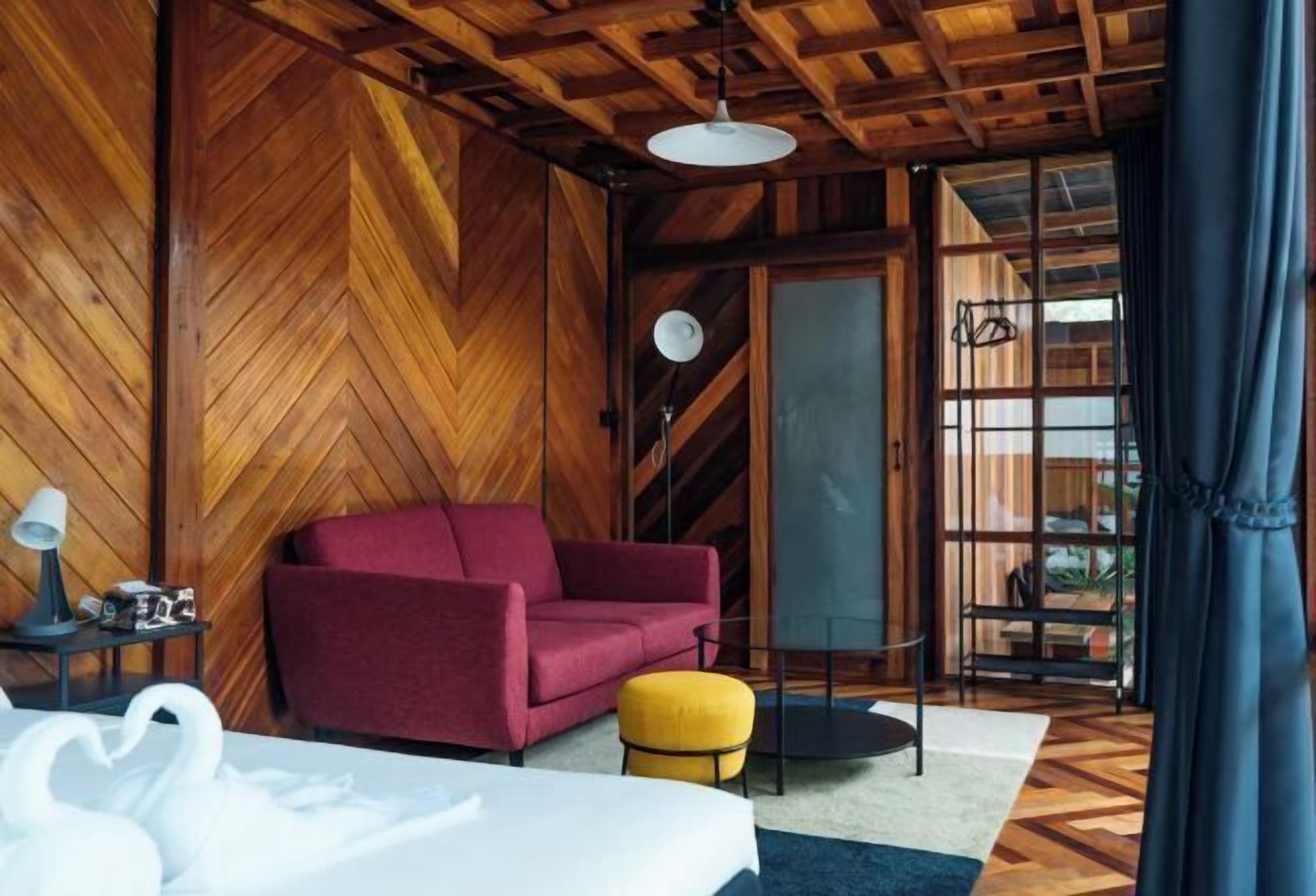 A room with a herringbone wooden wall, a red sofa, a yellow ottoman, a white bed, and a large window with curtains.