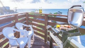 Un balcon avec un barbecue, des chaises en plastique blanc et une vue sur l’océan.