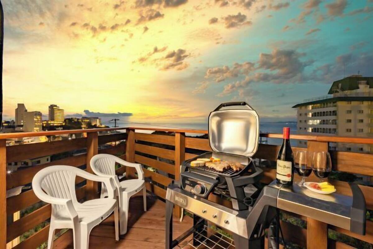 A rooftop terrace with a grill, two white plastic chairs, a bottle of wine, and a glass of red wine on a table.