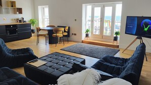 A modern living room with a black leather sofa, a black ottoman, and a large flat-screen TV. There is a dining area with a round table and chairs, and a kitchen area with a black countertop and cabinets. - Lavish Collections  (Great Yarmouth)