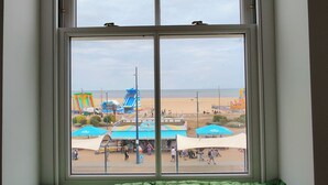 A window with a green cushioned bench below it, overlooking a beach with amusement park rides and umbrellas. - Lavish Collections  (Great Yarmouth)