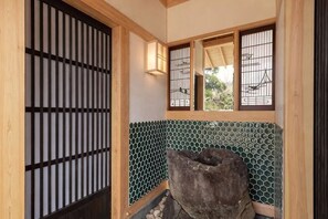 Une pièce de style japonais traditionnel, avec un sol recouvert de tatamis, un paravent en papier de riz (shoji), un bassin en pierre et une vue sur l’extérieur à travers une fenêtre ornée d’une grille métallique.