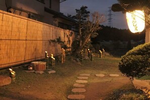 A pathway leading to a building with a bamboo fence, stone lanterns, and a small tree. - Tsubakihausu (Kozaki)