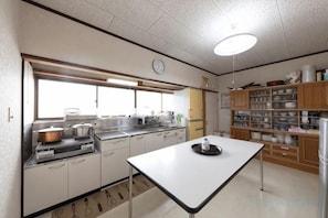 A kitchen with white cabinets, a stove, and a window. There is a clock on the wall and a shelf with dishes. - Tsubakihausu (Kozaki)