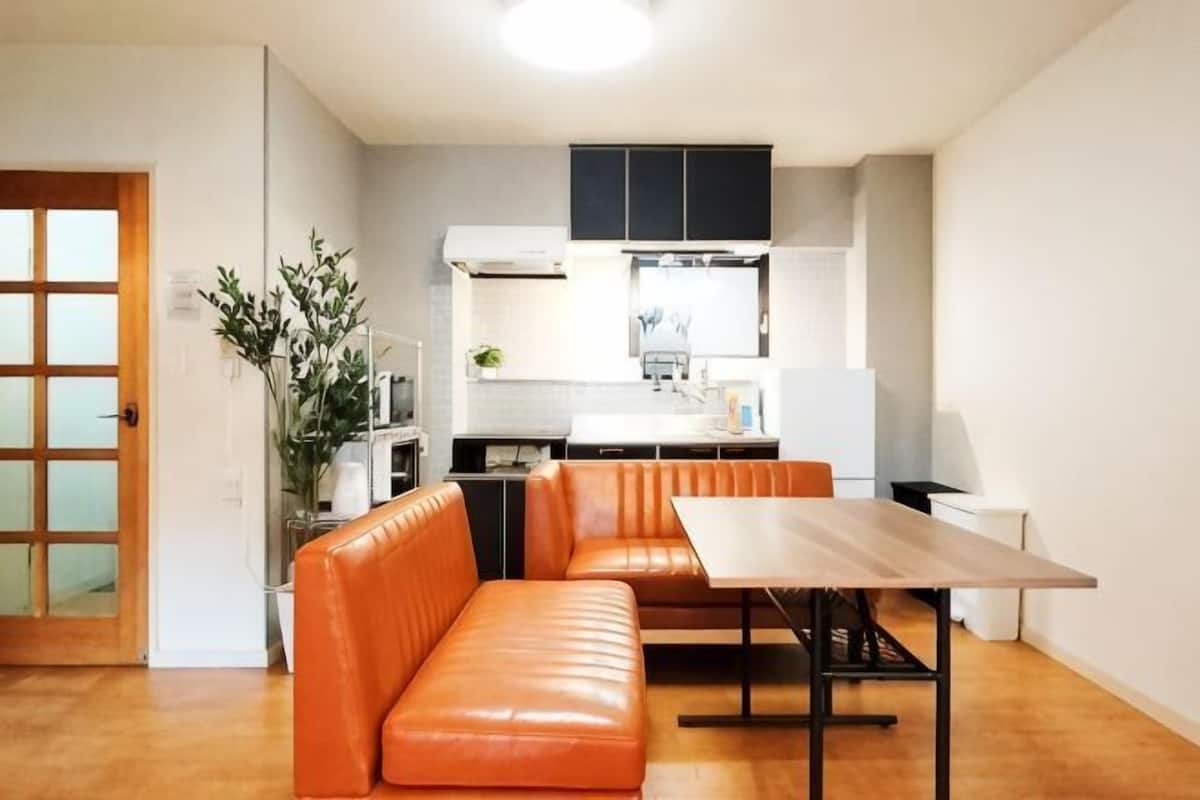 Ett kompakt bostadsutrymme med ett litet kök, en matplats med bord och orange sittmöbler samt ett trägolv.