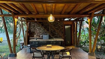 A covered outdoor dining area with a round table and chairs, a hanging light fixture, and a stone wall in the background.