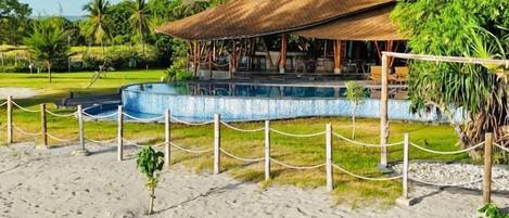 Un resort frente a la playa con piscina, un espacio de descanso cubierto y un camino de arena.