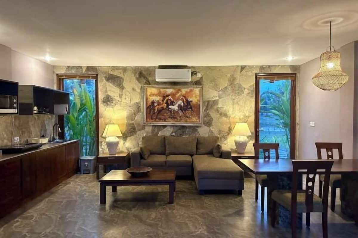 Una sala de estar con sofá, mesa de centro y comedor con sillas. Hay un cuadro en la pared, un televisor a la izquierda y una ventana con vistas a zonas verdes.