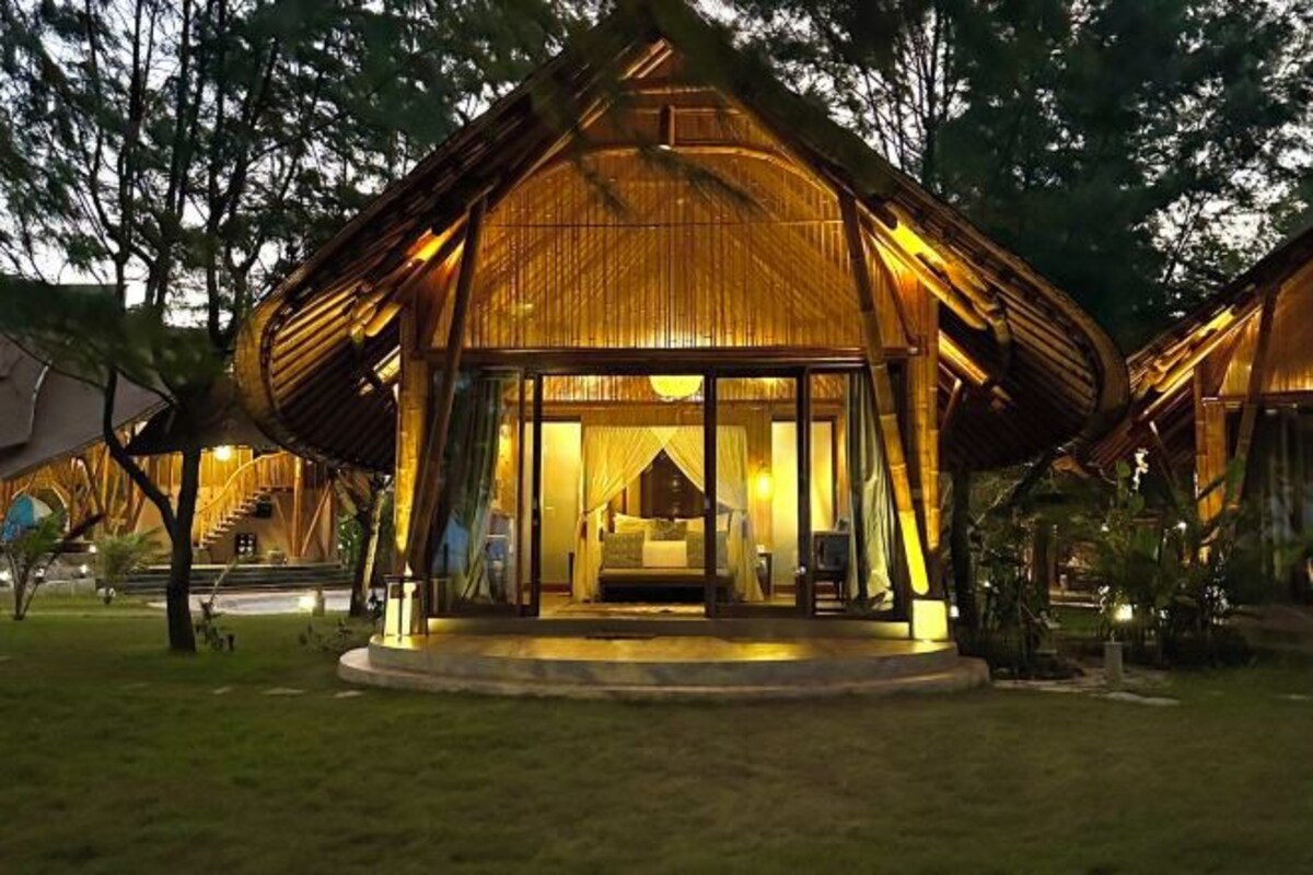 Une structure à toit de chaume avec une entrée en verre, un lit et un petit coin salon visible à l’intérieur.