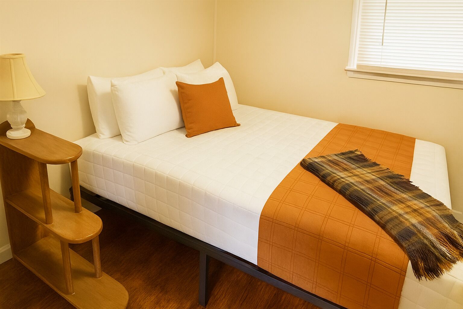 A single bed with white bedding and a plaid throw blanket, a wooden nightstand with a lamp, and a window with blinds.