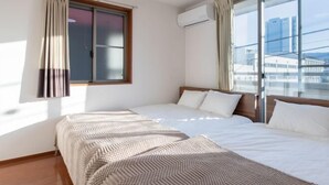 A hotel room with two beds, a large window with curtains, an air conditioning unit, and a view of buildings. - Residential Voyan Minamisunacho (Tokyo)