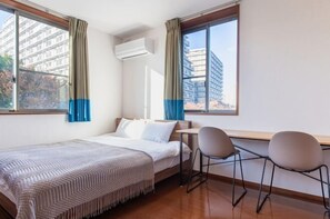 A hotel room with a bed, a desk, two chairs, an air conditioning unit, and large windows with curtains. - Residential Voyan Minamisunacho (Tokyo)