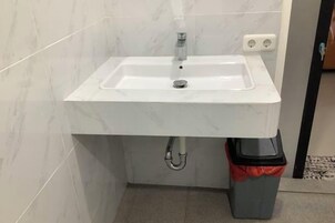 Un baño con lavamanos blanco, espejo y un cubo de basura.
