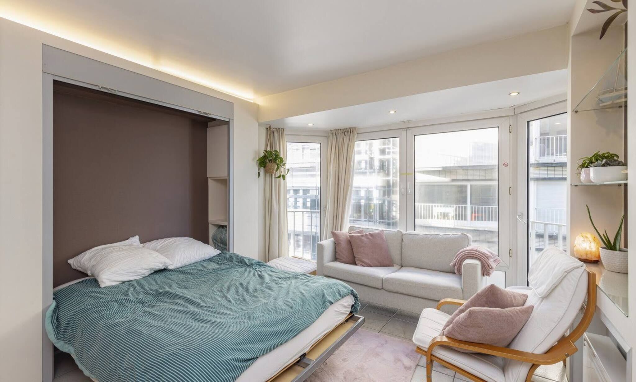 A modern bedroom with a bed, a sofa, and a chair. There are windows with a view of the city.