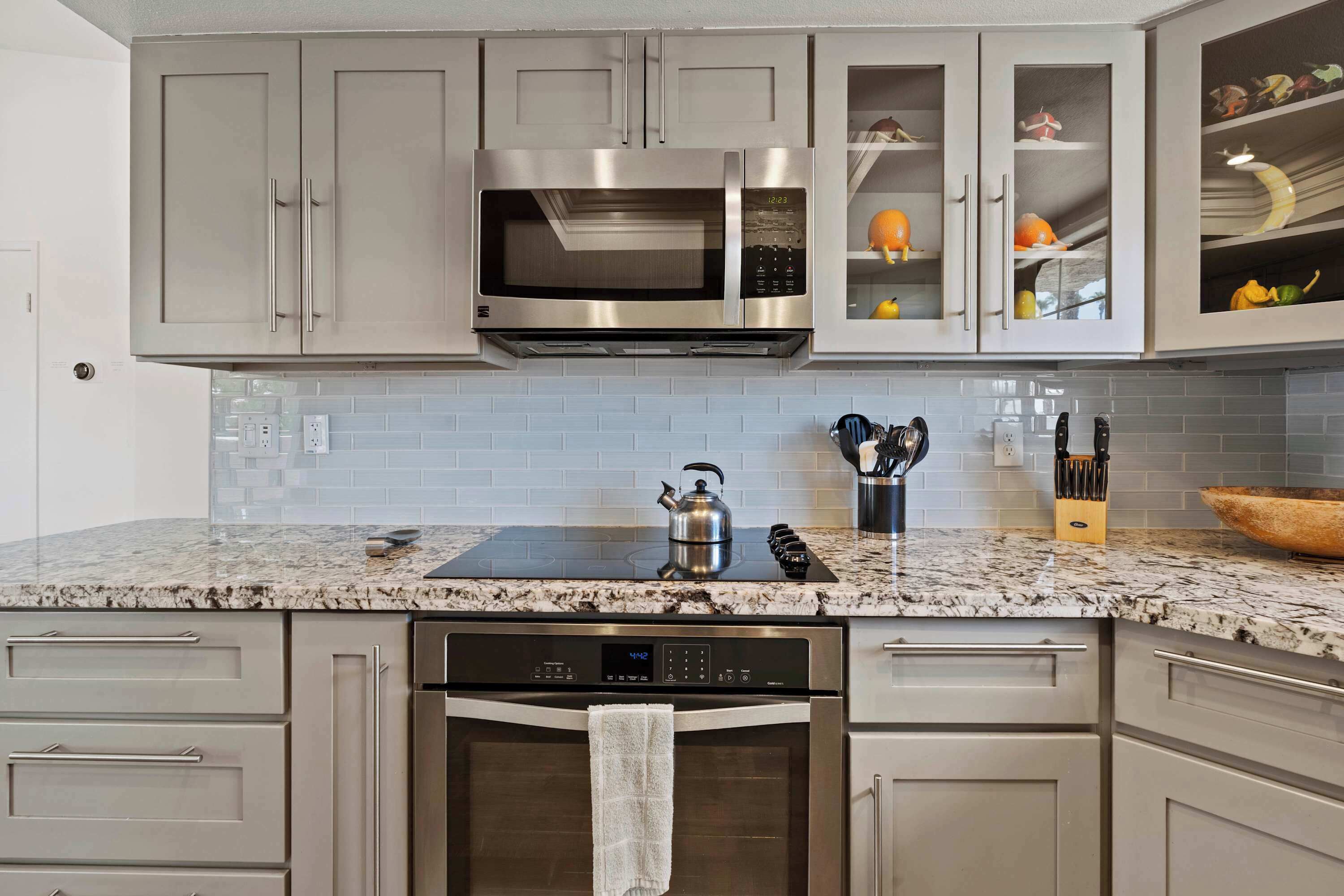 A modern kitchen with grey cabinets, a stainless steel microwave, a built-in oven, a kettle, and a knife block.