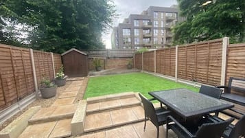 Ein Garten mit Terrassentisch und Stühlen, einer kleinen Gartenhütte und einem grünen Rasen.