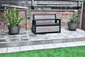 A bench with a metal frame and wooden slats, positioned on a stone-paved area with two potted plants on either side. - Modern Garden Retreat - Near Wfj, 3Br Ensuite & Parking (Watford)